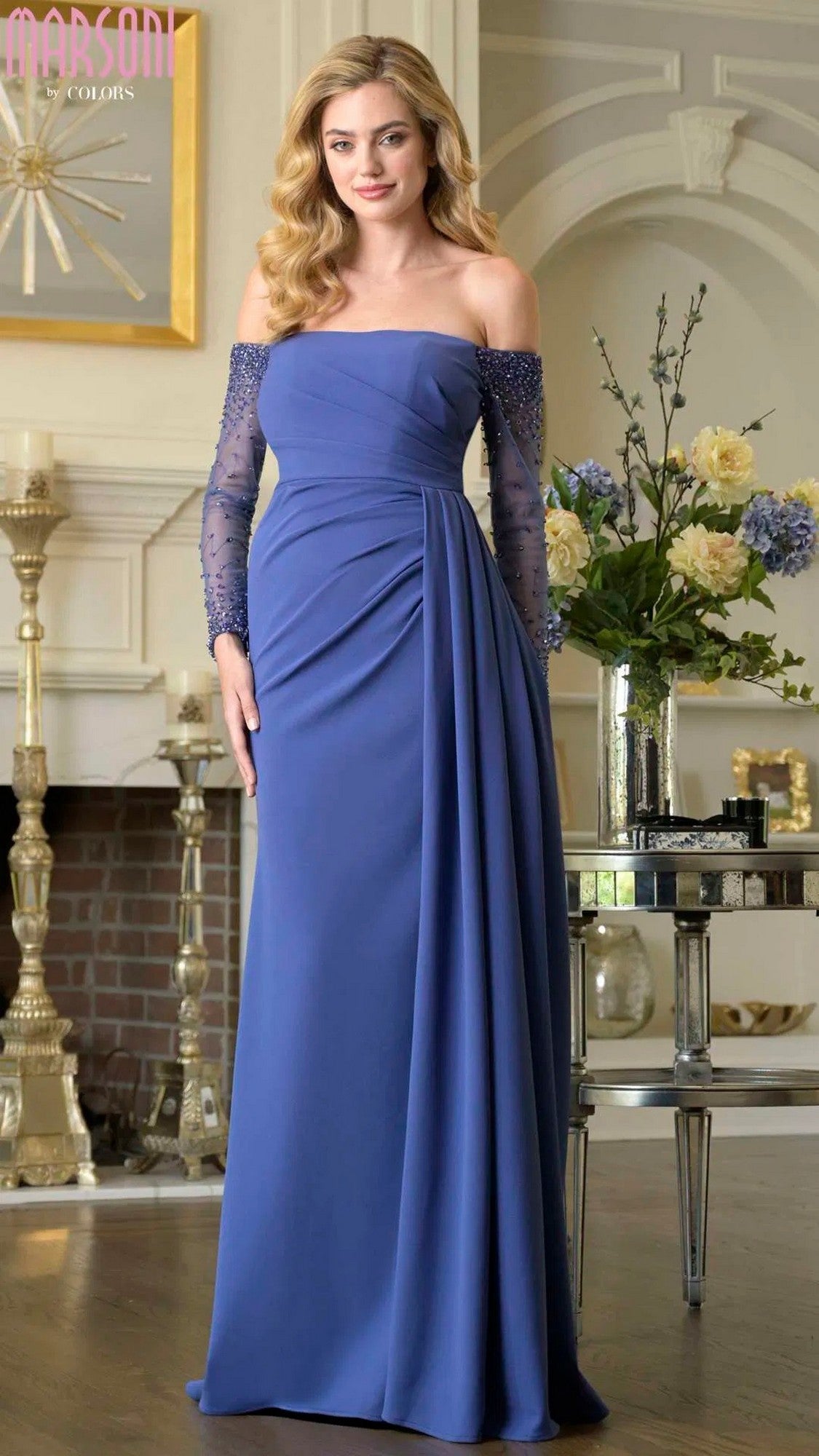 A woman wears the Marsoni M344 Long Formal Dress in Deep Royal with sheer long sleeves, standing in a decorated living room featuring a fireplace and floral arrangement.